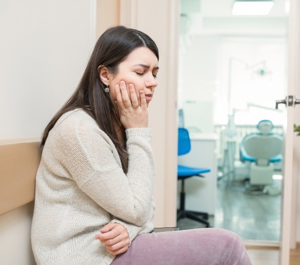 Woman sitting on bench in dental office holding her cheek in pain