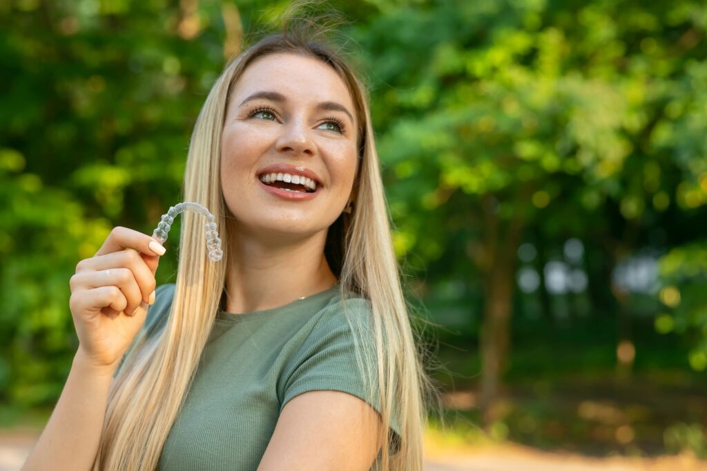 Woman in green shirt outside with trees holding clear aligner